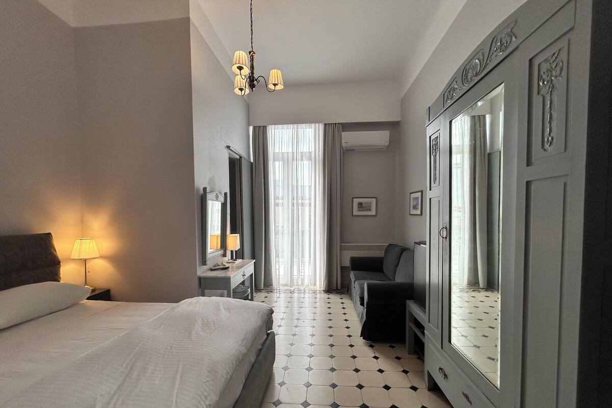 A hotel room with a bed, a desk, a chair, a large mirror, and a chandelier.