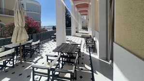 An outdoor balcony with tables and chairs, a covered area with an umbrella, and a view of the sea. - Tinion Urban Hotel (Tinos)