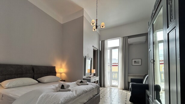 A hotel room with a large bed, a nightstand, a lamp, a dresser, and a view of the outside balcony. - Tinion Urban Hotel (Tinos)