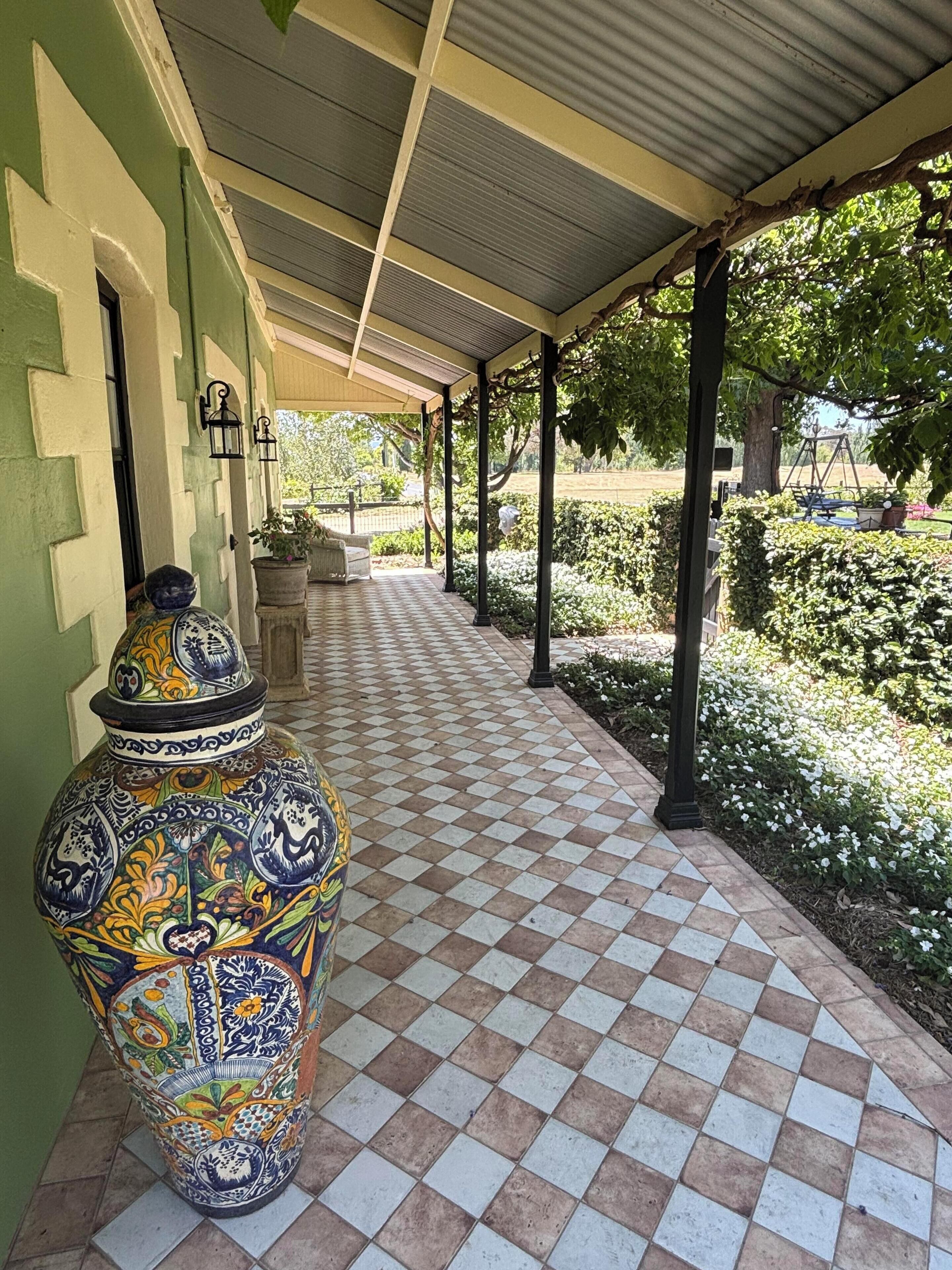 A covered walkway with a tiled floor, a decorative ceramic pot, and a garden area with white flowers and green shrubs.