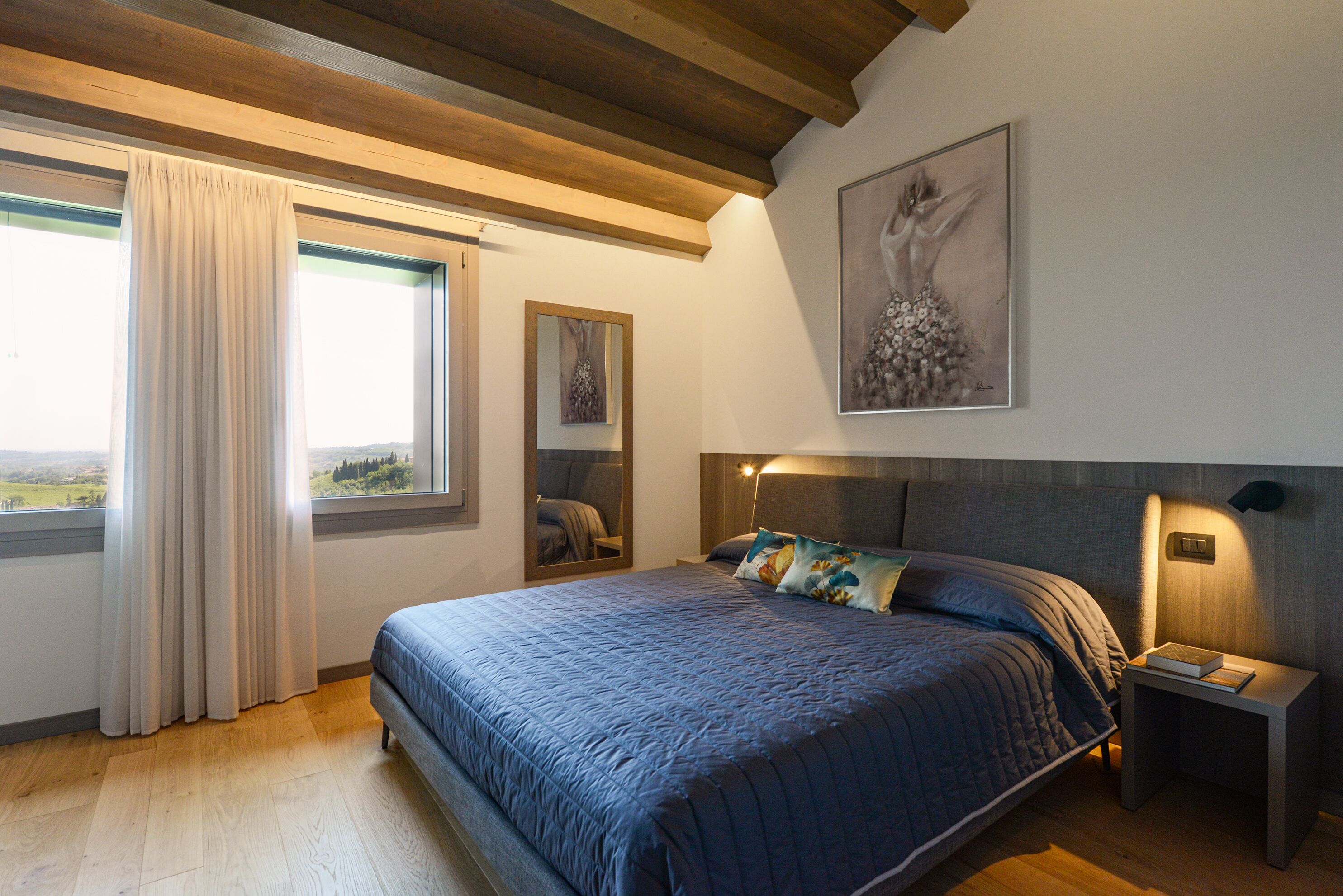 Een slaapkamer met een groot bed, een nachtkastje, een spiegel en een raam met gordijnen.