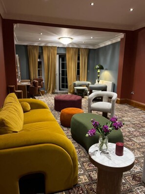 A modern hotel room with a yellow sofa, green ottoman, and a round side table with a vase of purple flowers.