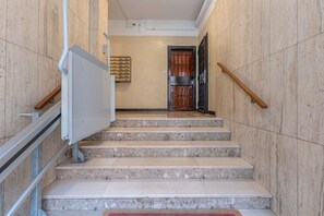 Un conjunto de escaleras de mármol con barandillas de metal, una puerta con reja metálica y una lámpara fijada a la pared.