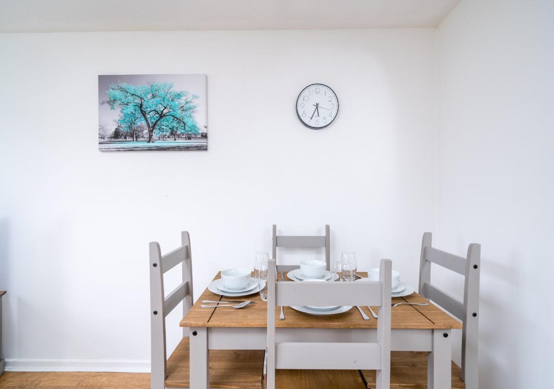 A dining area with a wooden table set for four, white chairs, a clock on the wall, and a framed picture above the table.