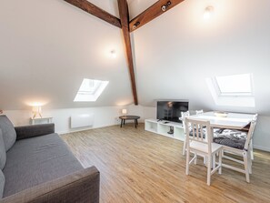 Un appartement moderne de style « loft », avec des poutres apparentes en bois, un lit en mezzanine, un coin repas avec une table et des chaises, un téléviseur et des fenêtres de toit.