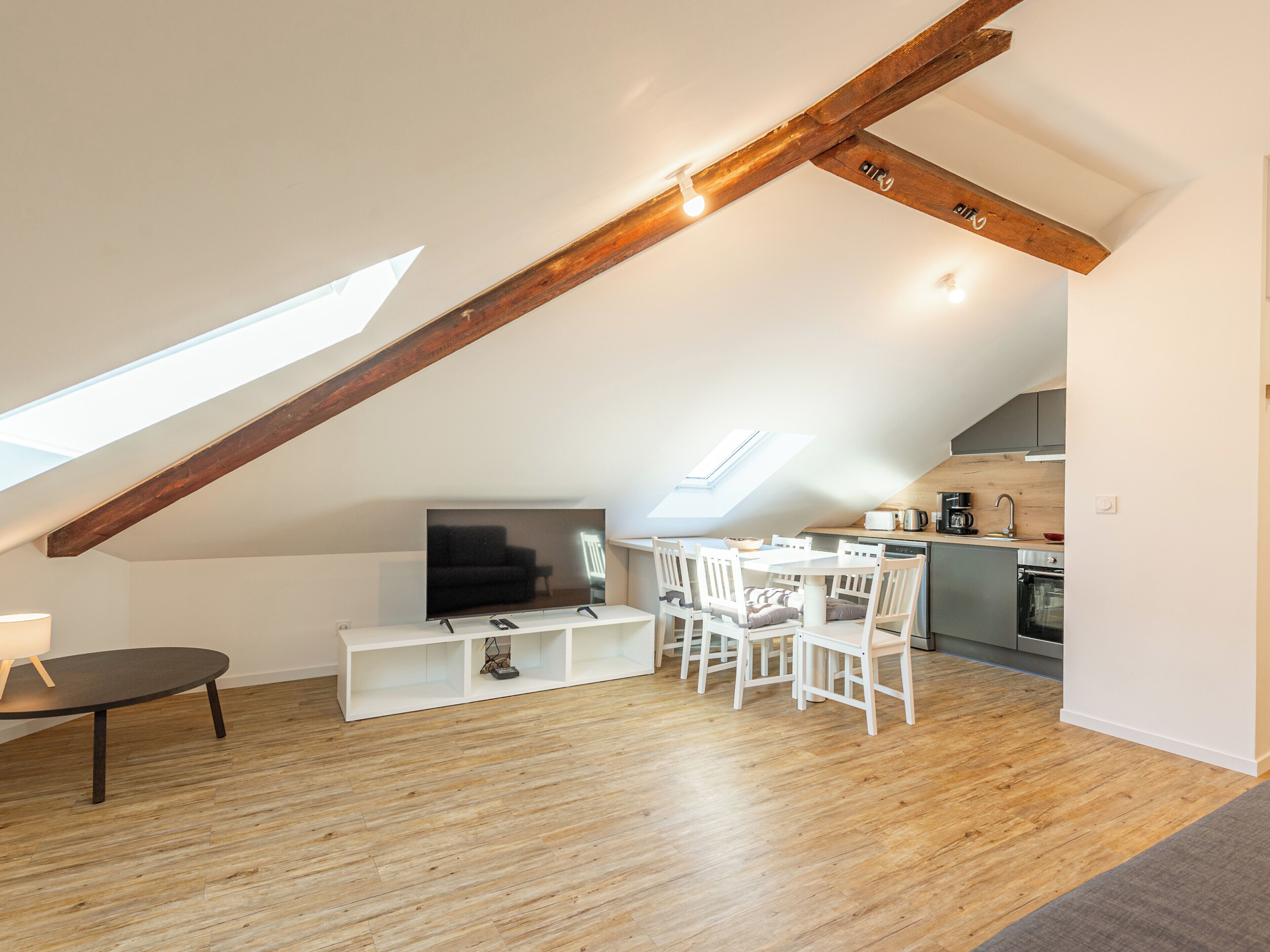 Un loft moderno con techo inclinado, vigas de madera, zona de comedor con sillas blancas, cocina con armarios oscuros y televisor de pantalla plana montado en un estante blanco.