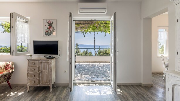 Een kamer met een televisie op een kast, een deur die uitkomt op een balkon met uitzicht op zee, en een plafondairconditioner.