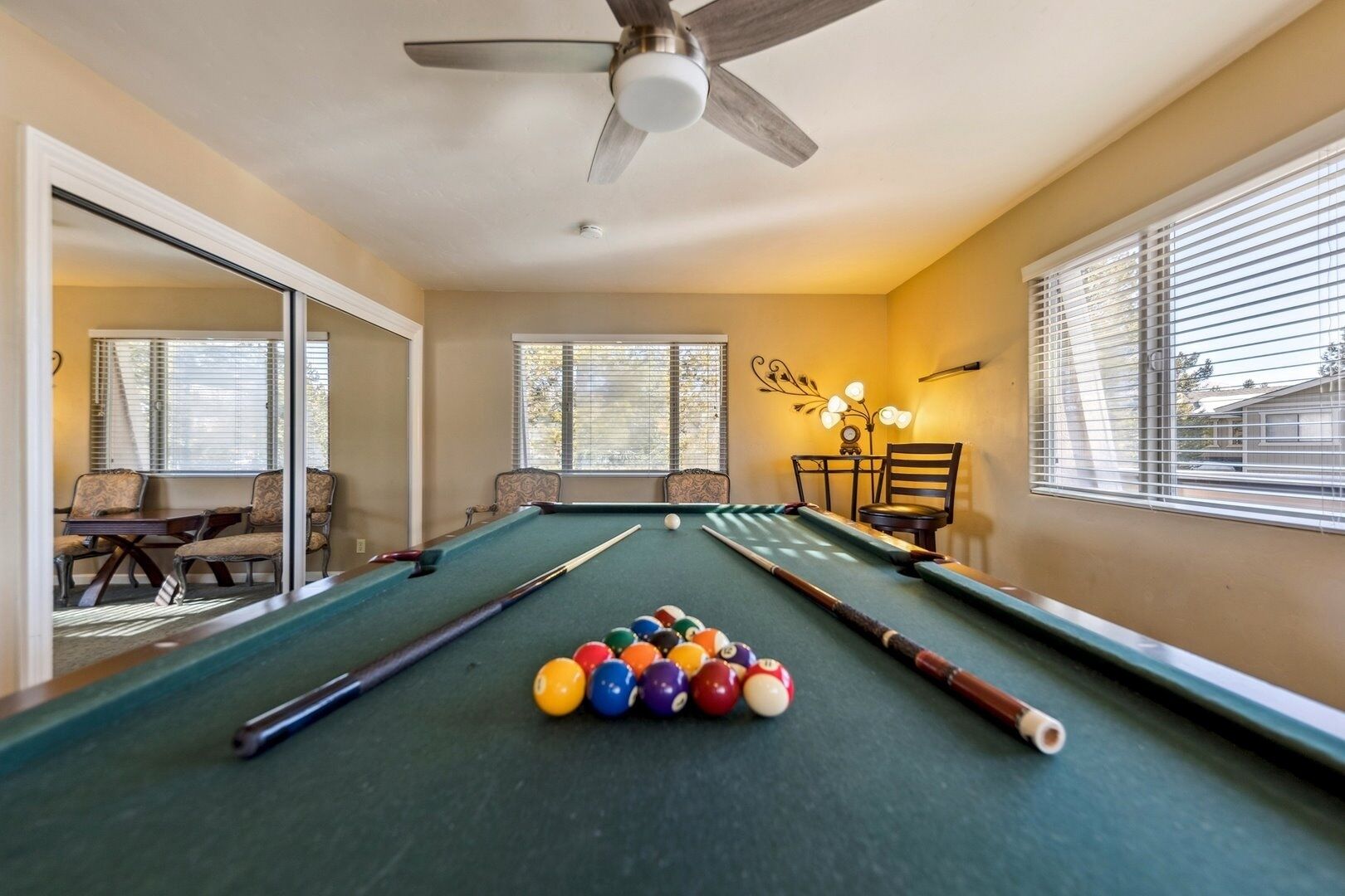 Une table de billard avec des billes, des chaises et un ventilateur de plafond dans une pièce dotée de fenêtres et d’un luminaire décoratif mural.