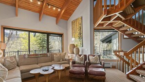 A spacious living room with a large sofa, armchairs, and a coffee table. A wooden staircase and large windows with a view of trees are visible. - Family-friendly Ski Escape With Hot Tub 4 Bedroom Condo (Park City)