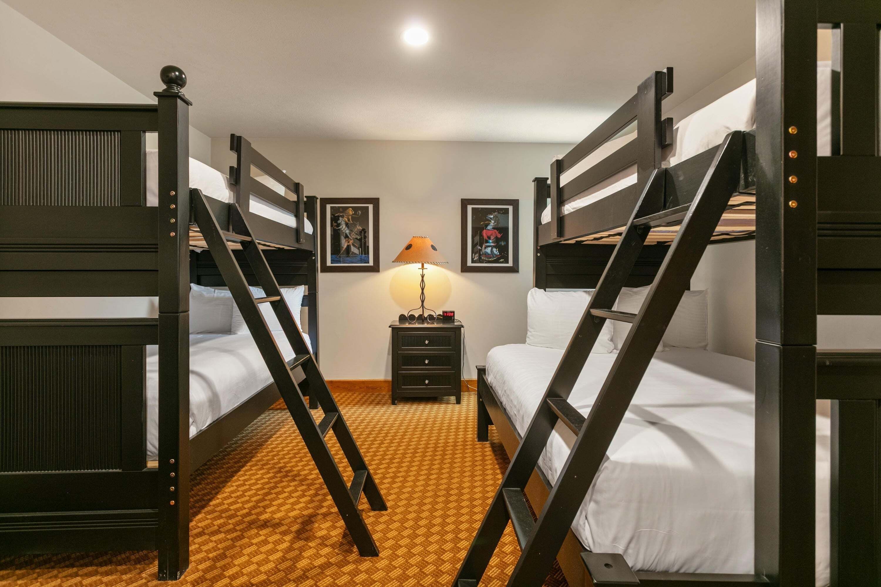 A room with bunk beds, a nightstand with a lamp, and framed pictures on the wall.