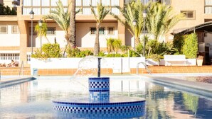 En swimmingpool med en kant av blått mosaik, omgiven av palmer och en byggnad med balkonger.