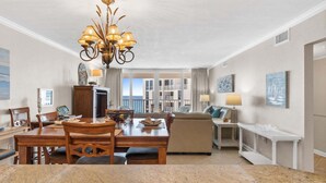 A spacious living room with a dining area, a chandelier, and a view of the balconies. - Shoreline Towers 1115 2 Bedroom Condo (Destin)