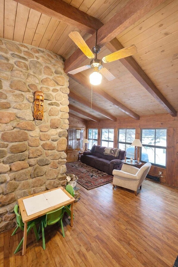 Um quarto acolhedor com parede de pedra, vigas de madeira no teto, ventilador de teto, sofá, cadeira, mesa e janela com vista.