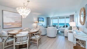 A spacious living area with a dining table, comfortable seating, a chandelier, and a large window offering a view of the sea. - Shoreline Towers 3095 2 Bedroom Condo (Destin)