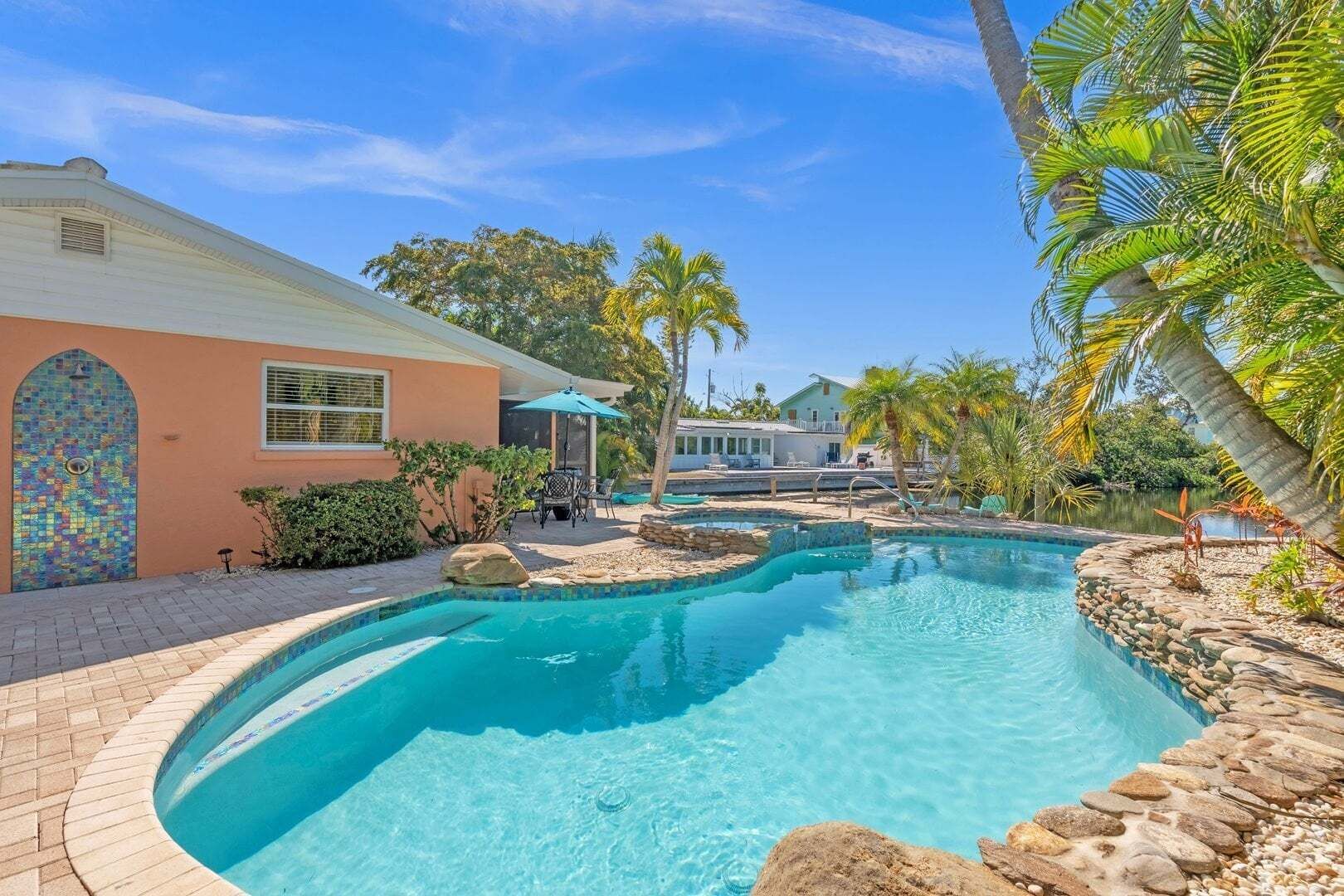 A swimming pool with a stone border, a patio area with outdoor furniture, and a house with a colorful mosaic door.