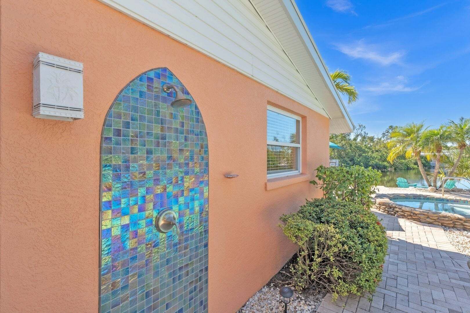 A house with a mosaic door, a small window, and a pool area with chairs and palm trees.