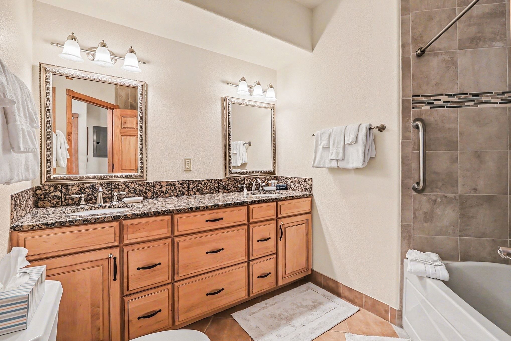 A bathroom with a double vanity, a large mirror, a shower with a grab bar, and a tiled floor.