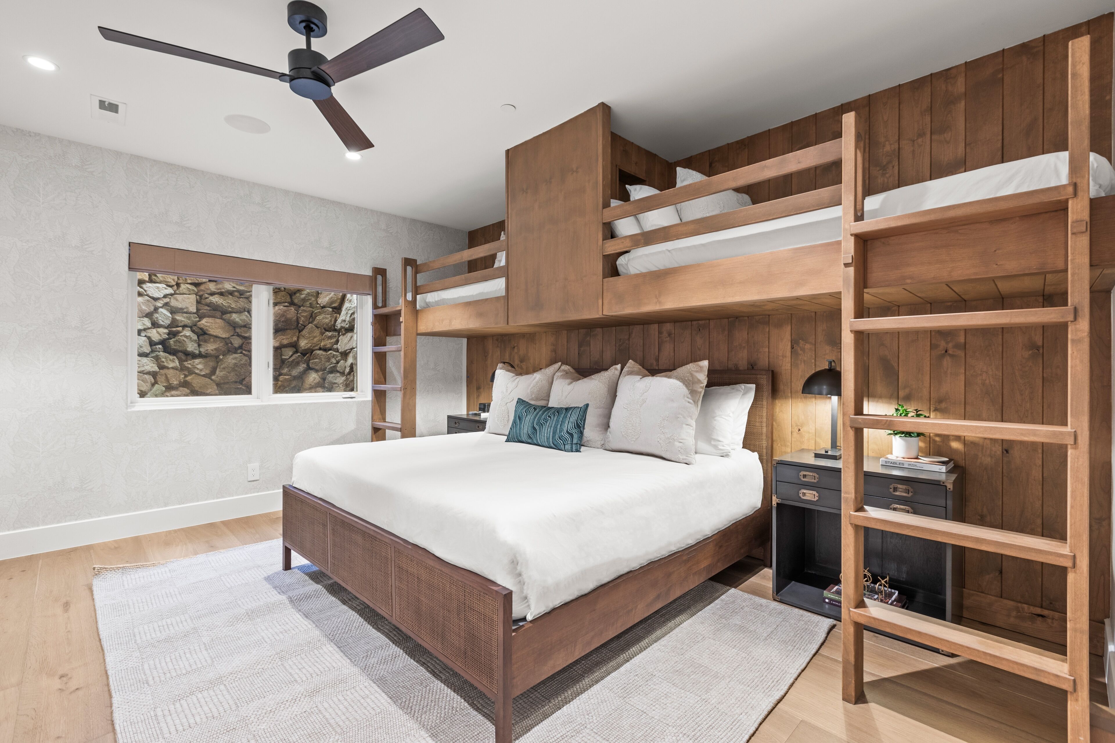 A bedroom with a bunk bed, a ceiling fan, a nightstand, and a window with a stone wall view.