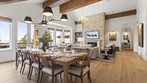 Uma sala de jantar espaçosa com uma mesa de madeira posta para seis pessoas, rodeada por cadeiras. Um candelabro está suspenso acima e uma lareira de pedra é visível ao fundo.