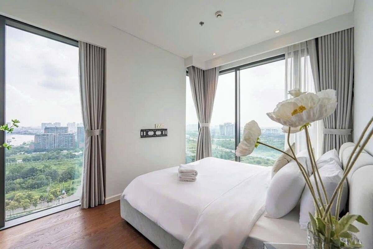 Quarto de hotel moderno com uma cama grande, roupa de cama branca e vista para a paisagem urbana através de janelas do chão ao teto.