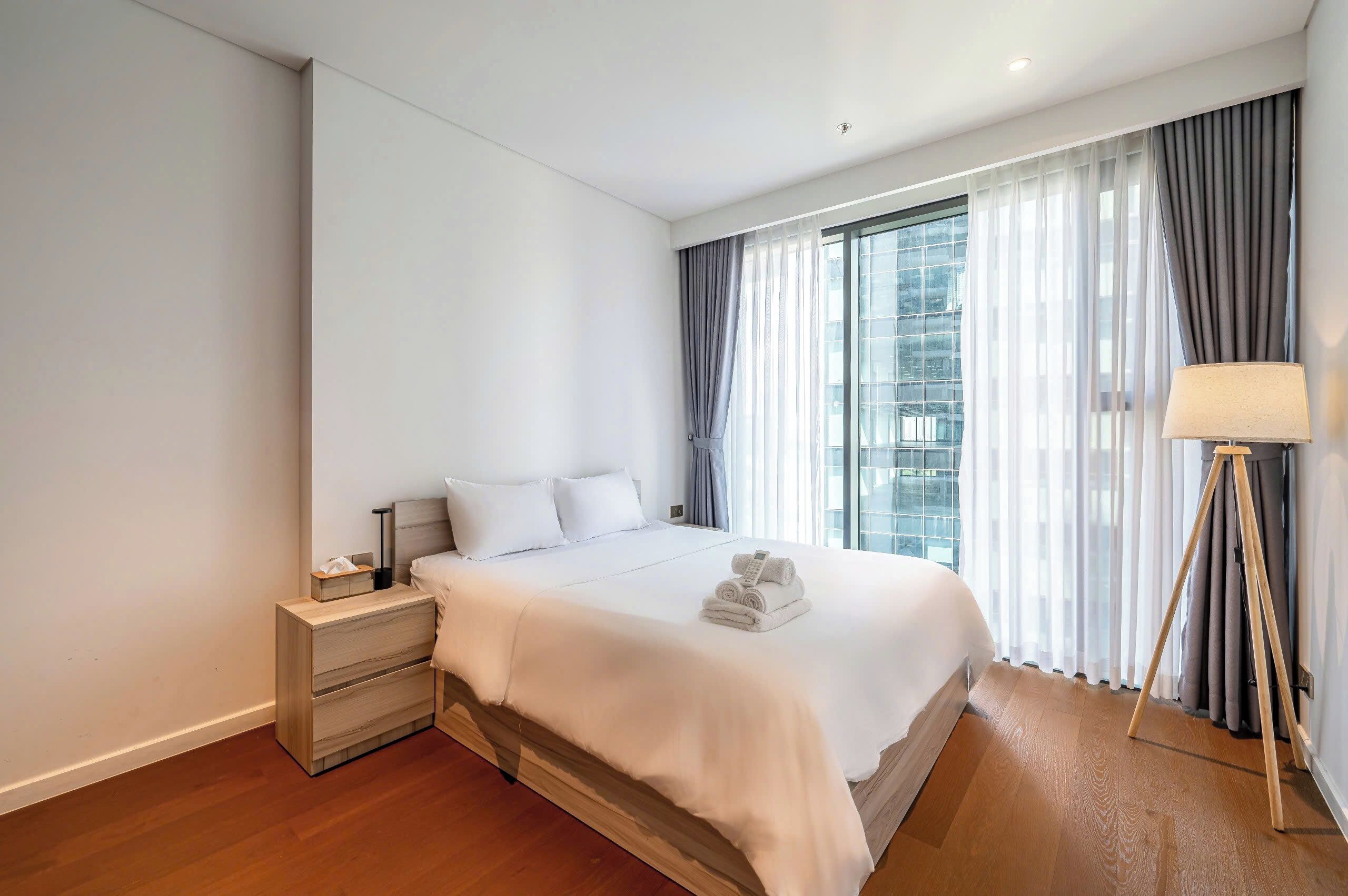 A hotel room with a large bed, a bedside table with a lamp, and a view of the city through sheer curtains.