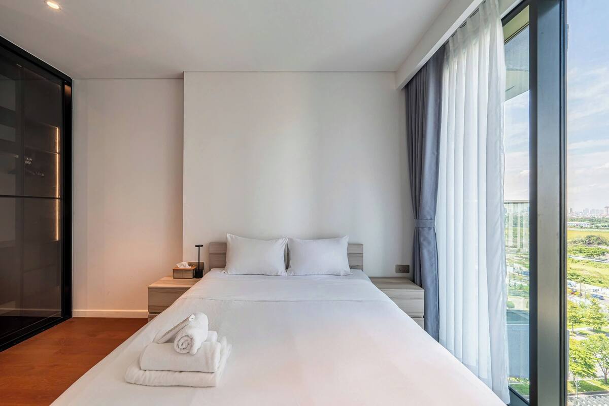Quarto de hotel moderno com uma cama grande, uma mesa de cabeceira, um armário de vidro e vista para a cidade pela janela.
