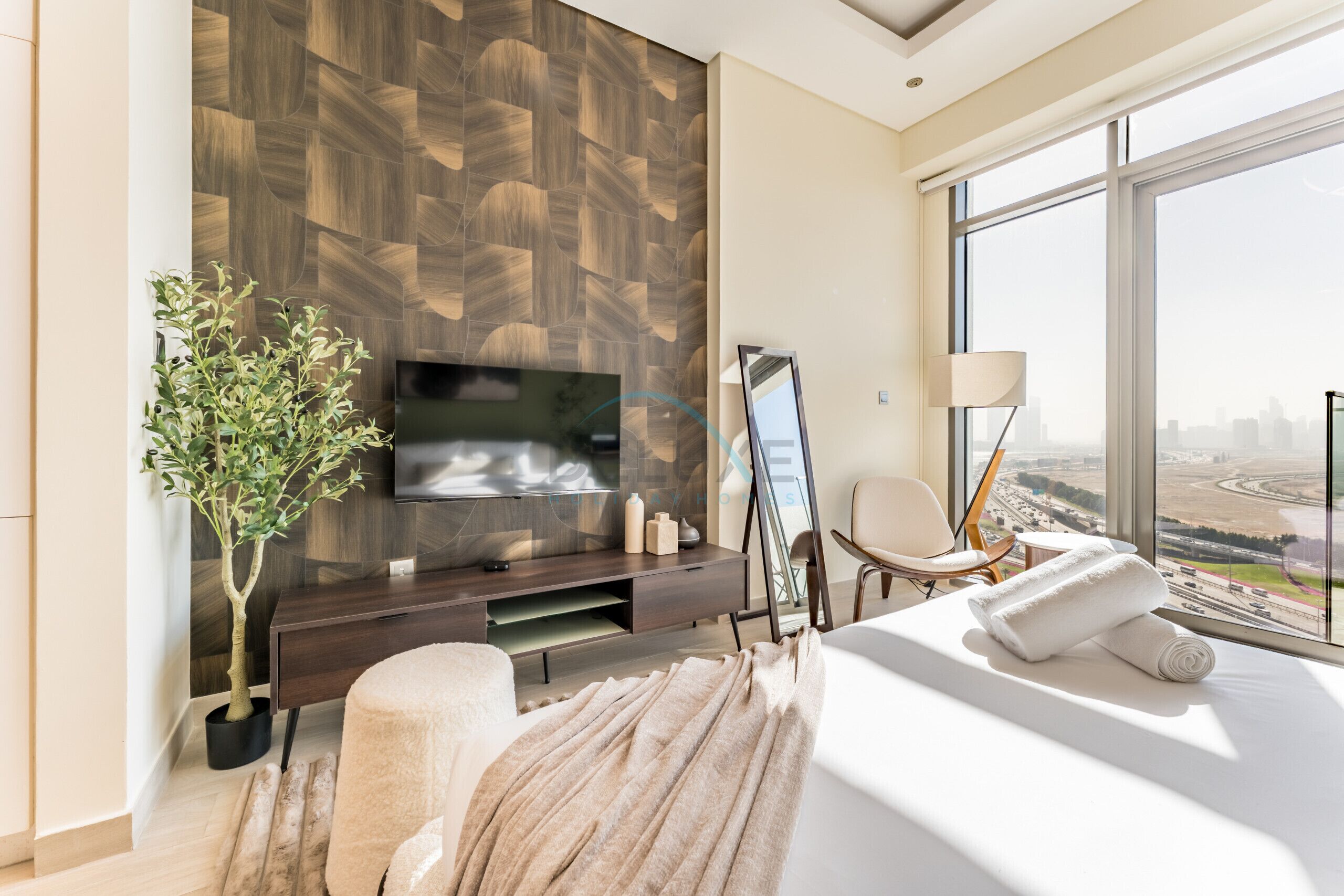 A modern hotel room with a large TV, a plant, a sofa, and a view of the city.