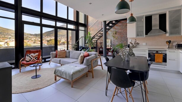 A spacious living area with a large window overlooking a cityscape, a modern kitchen, and a dining area with a table and chairs. - City Center Mountain Views The Piazza (Cape Town)