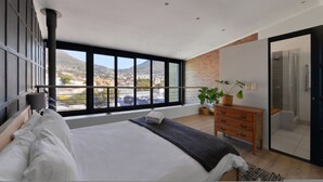A modern bedroom with a large bed, a dresser, a potted plant, and a view of a cityscape through large windows. - City Center Mountain Views The Piazza (Cape Town)