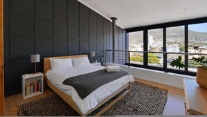 A modern bedroom with a large bed, a nightstand with books, a window with a view of the city, and a potted plant. - City Center Mountain Views The Piazza (Cape Town)