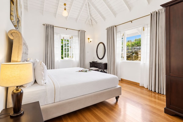 Une chambre à coucher avec un grand lit, une table de chevet avec une lampe, une commode, un miroir et une fenêtre avec des rideaux.