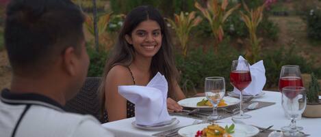 A dining table set outdoors with plates of food, glasses of red wine, and a white tablecloth.