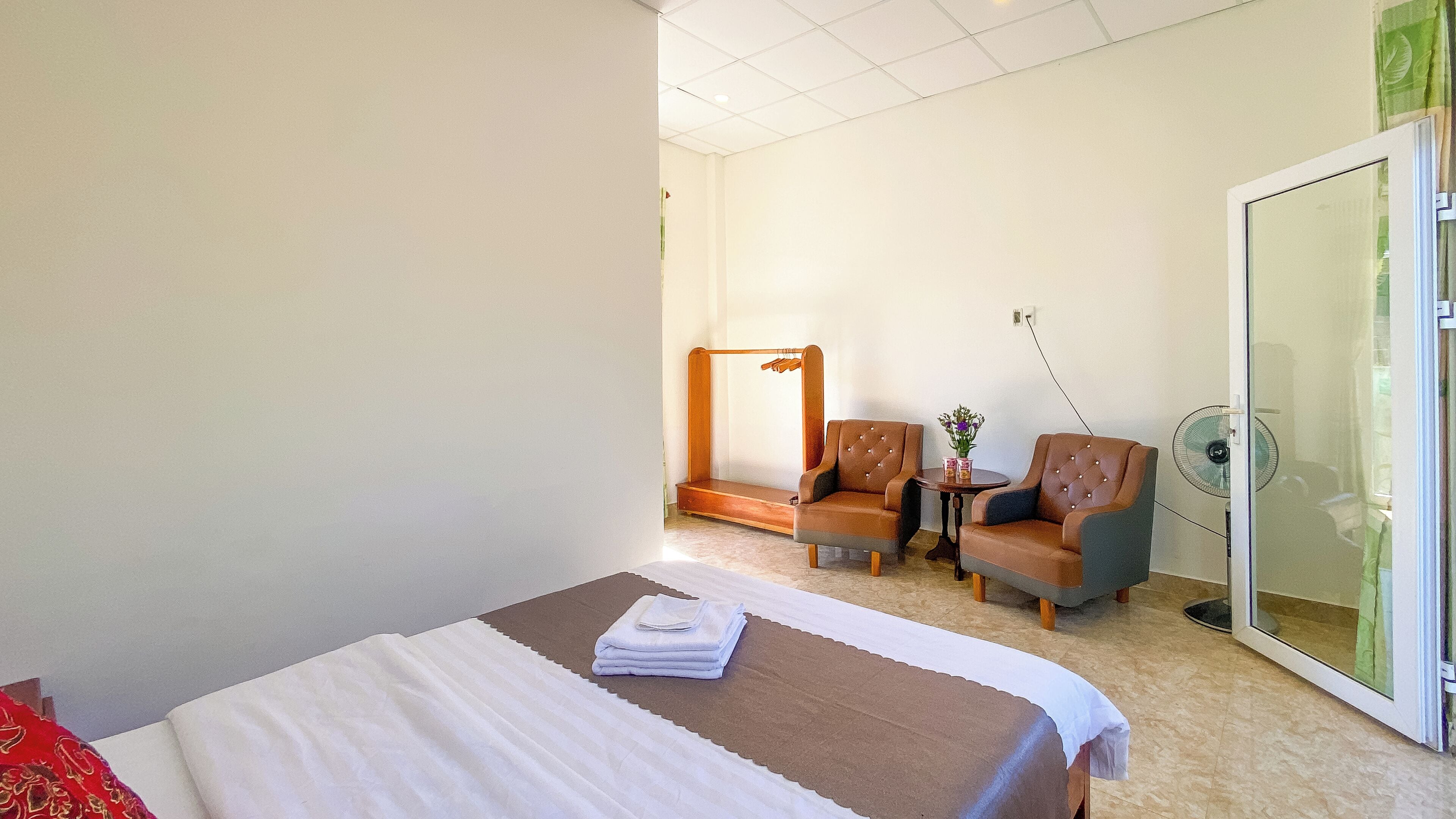 A hotel room with a bed, two armchairs, a small table with a vase, and a sliding glass door.
