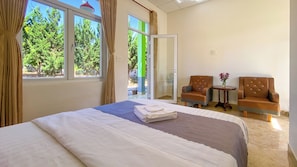 A hotel room with a bed, two armchairs, a small table with a vase, and a sliding glass door leading to an outdoor area with greenery.