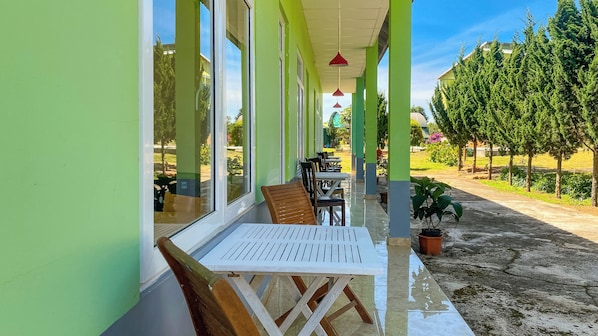 A covered outdoor seating area with tables and chairs, a potted plant, and a view of a garden and trees.