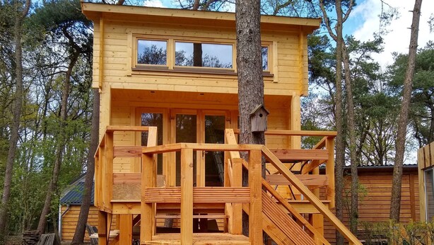 Una cabaña de madera con balcón y un nivel superior con ventanas, rodeada de árboles.