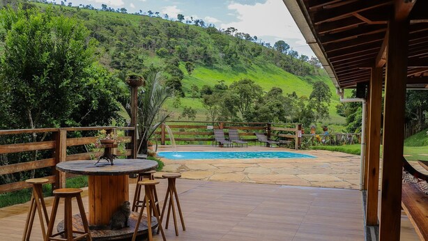 A wooden deck with a round table and stools, a pool, and a view of a green hillside. - S tio Toca dos Gatos com Piscina e Churrasqueira (Sabinópolis)