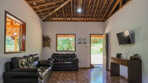 A spacious living room with a wooden ceiling, two black leather sofas, a wooden desk with a flat-screen TV, and large windows offering a view of the outdoors. - S tio Toca dos Gatos com Piscina e Churrasqueira (Sabinópolis)