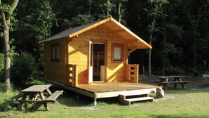 Una cabaña de madera con una plataforma elevada y la puerta abierta, rodeada de árboles.