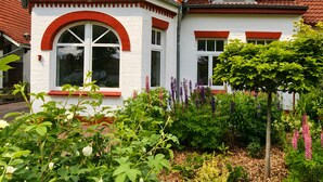 Une maison à deux étages, avec un toit recouvert de tuiles rouges, des murs blancs et un jardin où poussent différentes espèces de plantes.