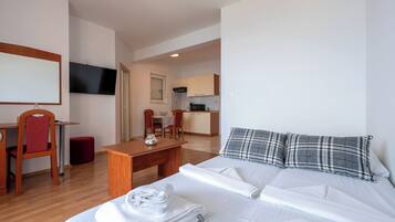 A hotel room with a bed, a desk, a chair, a TV, and a kitchenette in the background.