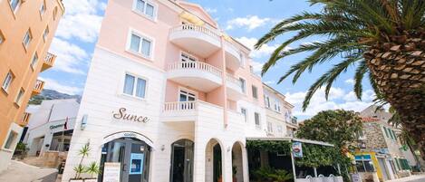 A multi-story hotel with a pink and white facade, a central entrance with an archway, and a balcony on the upper floor.