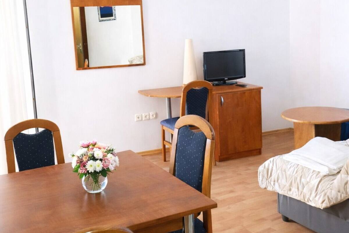 A hotel room with a wooden dining table, two chairs, a small cabinet with a television, a bed, and a mirror on the wall.