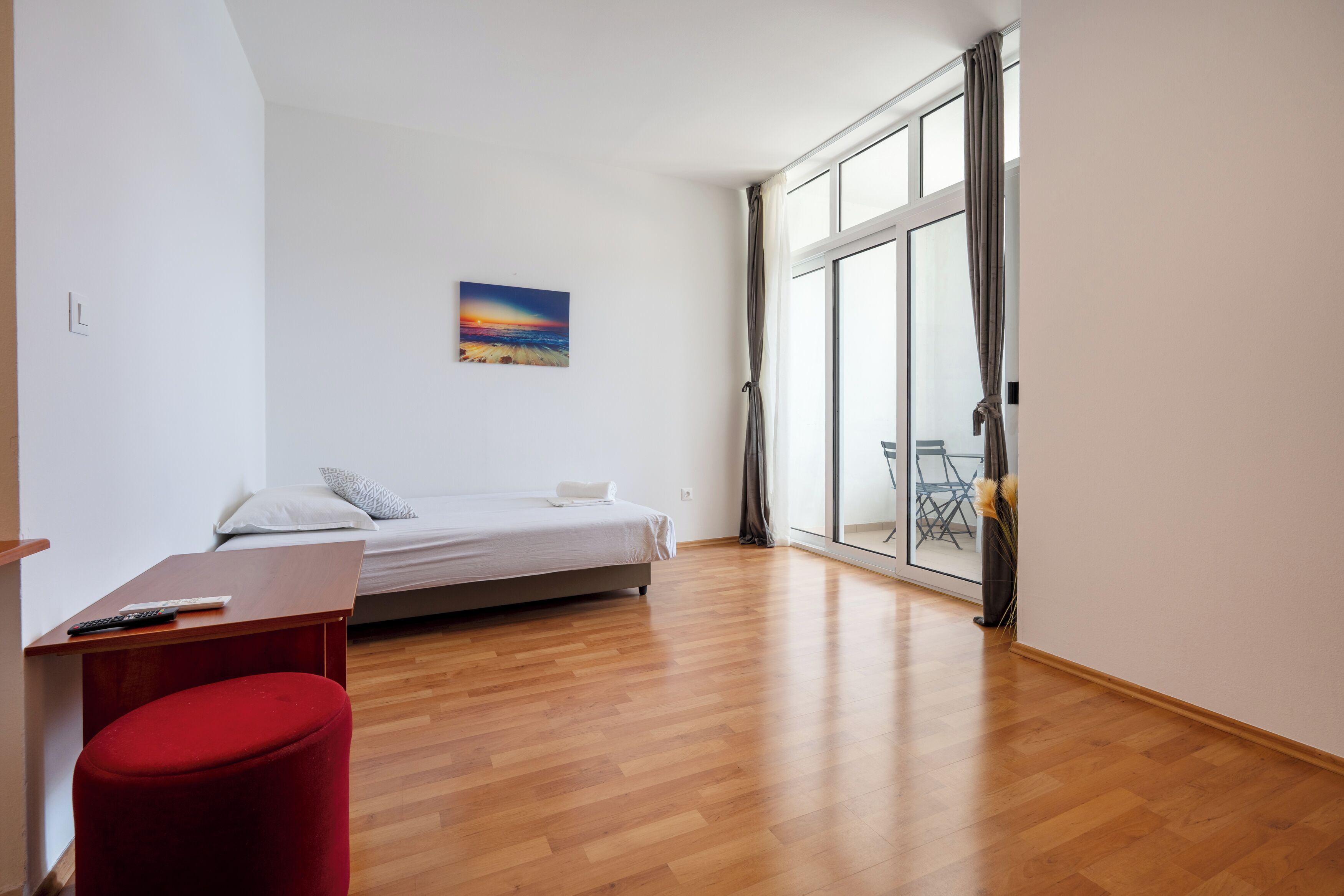 A bedroom with a bed, a red ottoman, a painting, and a view through a large window.