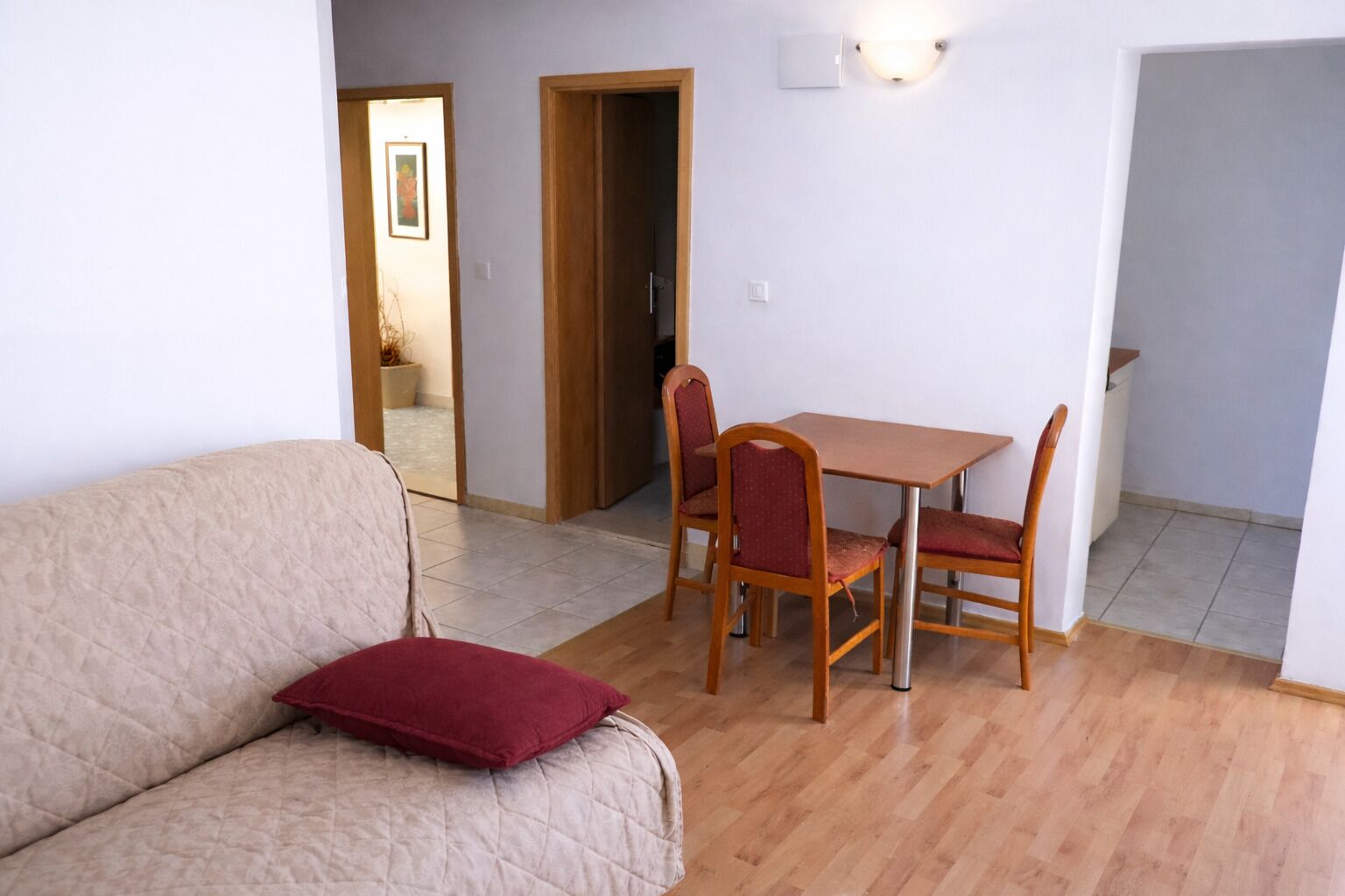 Ein Wohnzimmer mit einem beigen Sofa, einem roten Kissen, einem Holzesstisch mit vier Stühlen und einer Wandleuchte.