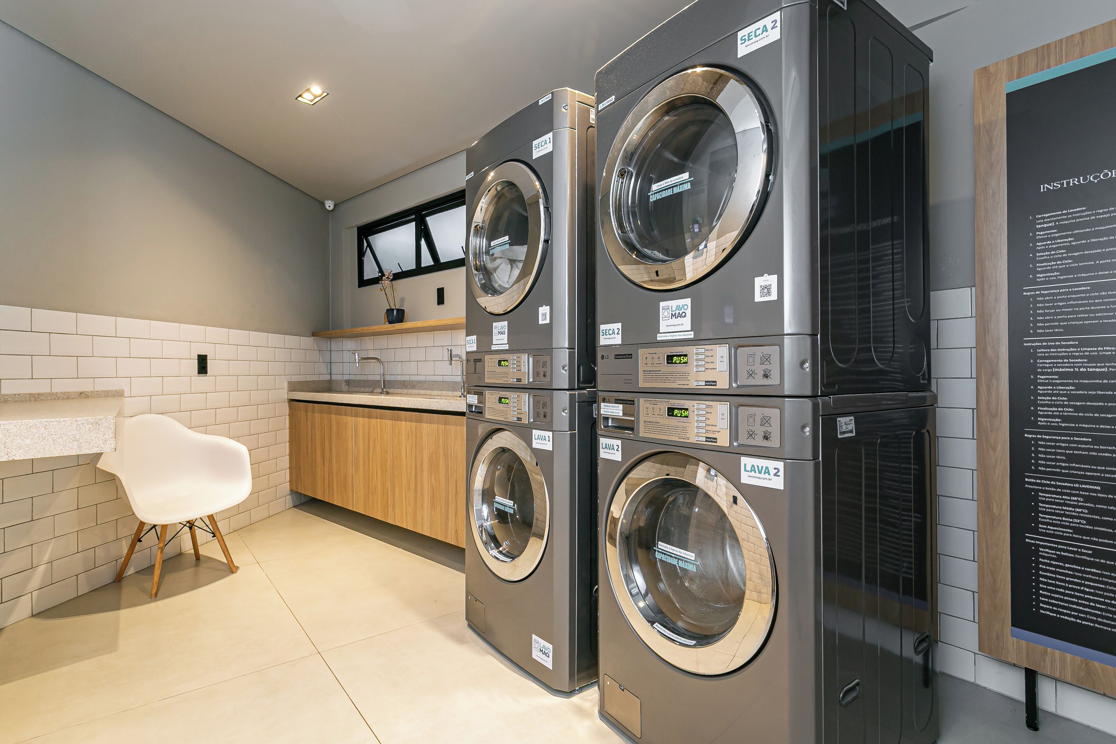 A modern laundry room with two stacked front-loading washing machines, a wooden cabinet, a white chair, and a wall-mounted instruction panel.