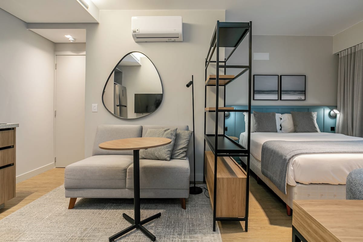 A modern hotel room with a sofa, a small round table, a bed with a headboard, a mirror, and a wall-mounted air conditioner.