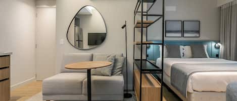A modern hotel room with a sofa, a small round table, a bed with a headboard, a mirror, and a wall-mounted air conditioner.