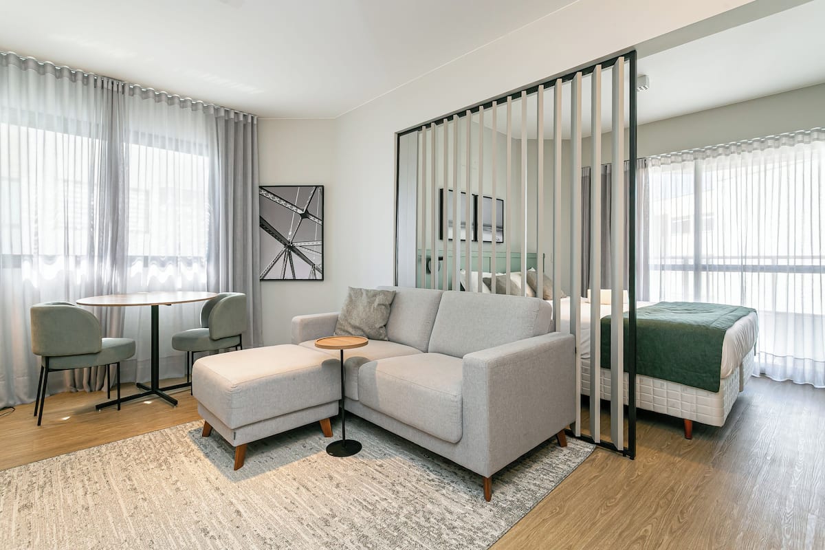 A modern, well-lit room with a grey sofa, a small round table, and a bed with a green blanket.