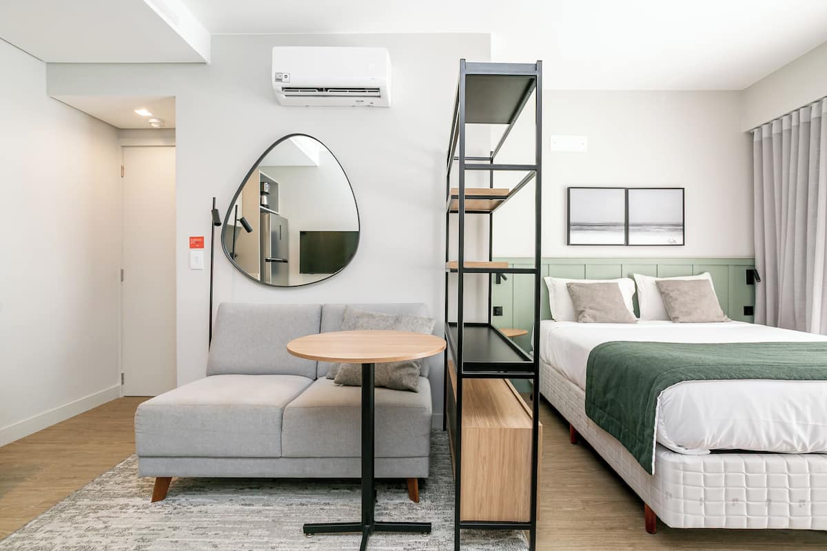 A modern hotel room with a sofa, a round table, a bed with a green blanket, a mirror, and a shelf unit.